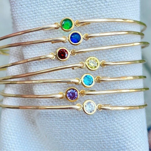 Dainty birthstone bangle featuring sparkling CZ diamonds, shown on a white fabric background 14k gold filled bangles, with red, blue, green, yellow, aqua, purple, clear focal cz center.
